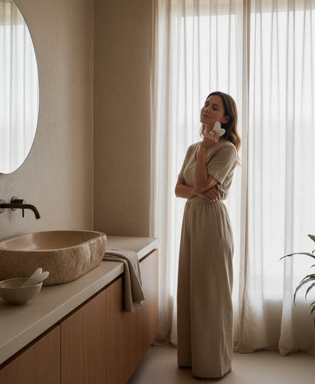 Vrouw gebruikt een witte jade gua sha tool tijdens een avond selfcare ritueel in een warme, minimalistische badkamer.