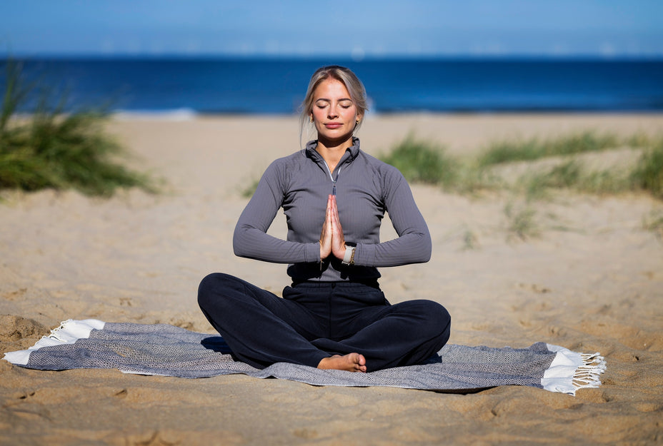 Yogadocent Muriel zittend in meditatie op het strand van Scheveningen, met de zee op de achtergrond.